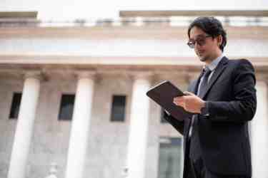 Lawyer in a dark suit and glasses reading a tablet in front of a columned, courthouse-style building